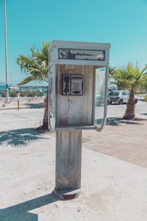 An outdoor cardphone booth with a weathered appearance stands on a paved area. There are palm trees and a road with cars in the background under a clear blue sky. The sign above the booth displays text in both Greek and English.