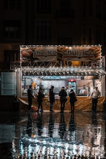 A group of people stand in line at a churro kiosk, which is adorned with festive lights. The kiosk is open for business in a dimly lit street, with reflections visible on the wet pavement. Inside the kiosk, two employees are preparing and serving food.