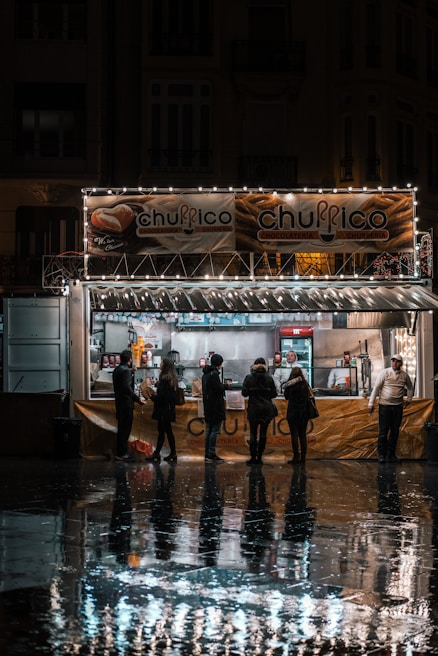 A group of people stand in line at a churro kiosk, which is adorned with festive lights. The kiosk is open for business in a dimly lit street, with reflections visible on the wet pavement. Inside the kiosk, two employees are preparing and serving food.