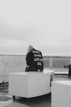 A person wearing a hoodie sits on a white, curved bench in an outdoor setting. The back of the hoodie has motivational text that reads 'WORK HARD DREAM BIG.' The scene is monochrome, creating a contemplative and minimalistic atmosphere. In the background, there is a barrier, suggesting this might be a rooftop or a balcony with a view.