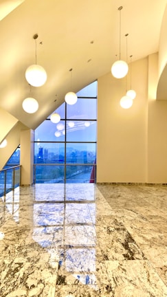 A modern interior space features large spherical pendant lights hanging from a high ceiling. The floor is made of polished stone tiles with a marbled pattern. A large floor-to-ceiling window allows natural light to enter, offering a view of the cityscape outside.