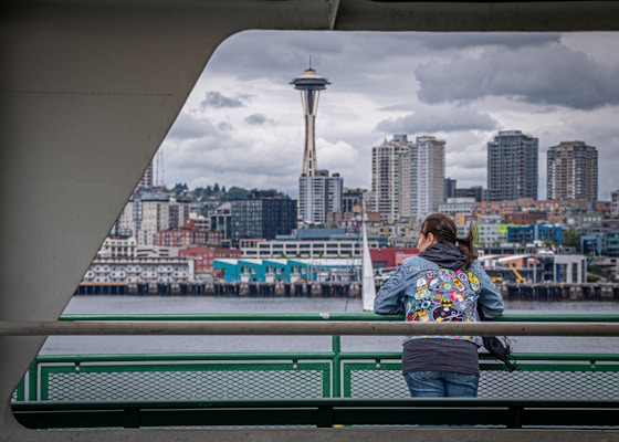 A person wearing a colorful jacket with patches stands on a ferry deck, gazing at the Seattle skyline. The iconic Space Needle and city buildings form a distinctive urban backdrop under a cloudy, overcast sky.