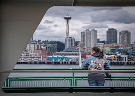 A person wearing a colorful jacket with patches stands on a ferry deck, gazing at the Seattle skyline. The iconic Space Needle and city buildings form a distinctive urban backdrop under a cloudy, overcast sky.
