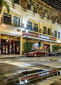 A street view featuring a multi-story building with warm-colored balconies and lit string lights across the top. The ground level has a bright, neon-lit sign for a venue. Cars are parked along the street, and the pavement has a wet, reflective surface that mirrors the lights and storefront.