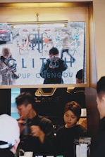 A cafe setting is depicted with people interacting at a counter. A reflective surface reveals a street scene with bicycles and pedestrians, showcasing a person taking a picture. The space appears to be bustling with activity, featuring a modern interior design with industrial elements.