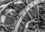 The image features an intricate view of a mechanical engine, showcasing various metal components, pipes, and bolts. The arrangement and complexity of the parts suggest a heavy-duty machine or vehicle engine. The texture and details of the metal surfaces are highlighted in black and white, emphasizing the industrial nature of the image.