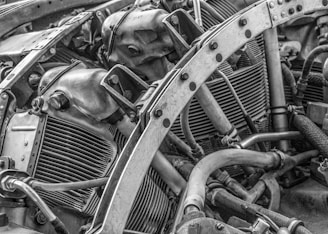 The image features an intricate view of a mechanical engine, showcasing various metal components, pipes, and bolts. The arrangement and complexity of the parts suggest a heavy-duty machine or vehicle engine. The texture and details of the metal surfaces are highlighted in black and white, emphasizing the industrial nature of the image.