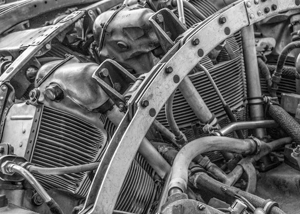 The image features an intricate view of a mechanical engine, showcasing various metal components, pipes, and bolts. The arrangement and complexity of the parts suggest a heavy-duty machine or vehicle engine. The texture and details of the metal surfaces are highlighted in black and white, emphasizing the industrial nature of the image.