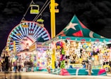 A vibrant carnival scene at night with a large, colorful Ferris wheel illuminated in the background. A brightly lit tent to the right displays an assortment of stuffed animals and prizes, attracting visitors. Overhead, a gondola ride with green cars adds to the festive atmosphere, while blurred figures of people walk throughout the scene, enhancing the lively and dynamic setting.