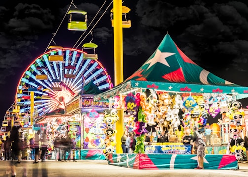 A vibrant carnival scene at night with a large, colorful Ferris wheel illuminated in the background. A brightly lit tent to the right displays an assortment of stuffed animals and prizes, attracting visitors. Overhead, a gondola ride with green cars adds to the festive atmosphere, while blurred figures of people walk throughout the scene, enhancing the lively and dynamic setting.
