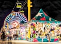 A vibrant carnival scene at night with a large, colorful Ferris wheel illuminated in the background. A brightly lit tent to the right displays an assortment of stuffed animals and prizes, attracting visitors. Overhead, a gondola ride with green cars adds to the festive atmosphere, while blurred figures of people walk throughout the scene, enhancing the lively and dynamic setting.