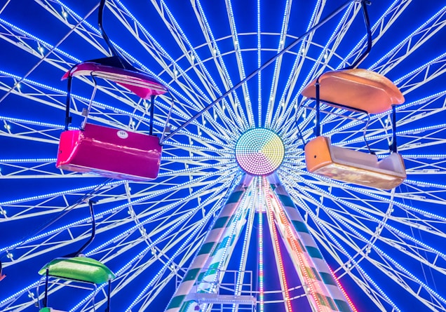 A vibrant ferris wheel with illuminated spokes creating a mesmerizing radial pattern against a deep blue sky. Colorful gondolas, including red, green, and orange, hang in the foreground, adding dynamic elements to the scene.