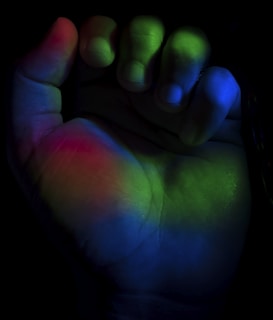 A human hand partially clenched, illuminated with a spectrum of colors including green, red, and blue. The light casts an ethereal glow across the palm and fingers, creating a striking contrast against the dark background.