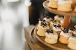 Small, individually-sized cakes with a light brown color and topped with white icing and blueberries are arranged on a wooden tiered stand. The cakes are adorned with small gold sprinkles and a garnish resembling dried fruit or herbs. The background is softly blurred, drawing focus to the cakes.