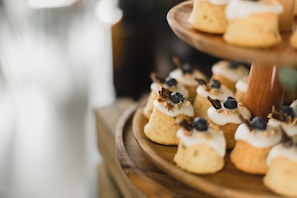 Small, individually-sized cakes with a light brown color and topped with white icing and blueberries are arranged on a wooden tiered stand. The cakes are adorned with small gold sprinkles and a garnish resembling dried fruit or herbs. The background is softly blurred, drawing focus to the cakes.