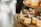 Small, individually-sized cakes with a light brown color and topped with white icing and blueberries are arranged on a wooden tiered stand. The cakes are adorned with small gold sprinkles and a garnish resembling dried fruit or herbs. The background is softly blurred, drawing focus to the cakes.