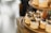 Small, individually-sized cakes with a light brown color and topped with white icing and blueberries are arranged on a wooden tiered stand. The cakes are adorned with small gold sprinkles and a garnish resembling dried fruit or herbs. The background is softly blurred, drawing focus to the cakes.