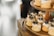 Small, individually-sized cakes with a light brown color and topped with white icing and blueberries are arranged on a wooden tiered stand. The cakes are adorned with small gold sprinkles and a garnish resembling dried fruit or herbs. The background is softly blurred, drawing focus to the cakes.