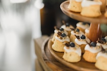 Small, individually-sized cakes with a light brown color and topped with white icing and blueberries are arranged on a wooden tiered stand. The cakes are adorned with small gold sprinkles and a garnish resembling dried fruit or herbs. The background is softly blurred, drawing focus to the cakes.