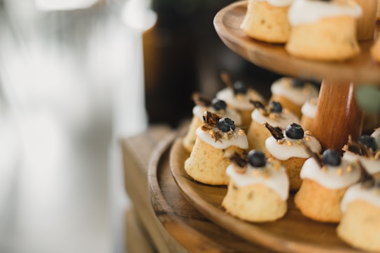 Small, individually-sized cakes with a light brown color and topped with white icing and blueberries are arranged on a wooden tiered stand. The cakes are adorned with small gold sprinkles and a garnish resembling dried fruit or herbs. The background is softly blurred, drawing focus to the cakes.