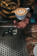 A person elegantly performs a trick or gesture with a cup of coffee, making it appear as if the cup is hovering above their hand. The cup contains a latte with intricate latte art on top. A metal countertop with circular perforations and a wooden surface nearby create an industrial and rustic environment.