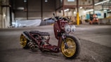 A custom motorcycle with a sleek design is parked inside an industrial warehouse. It features a maroon body, large dual headlights, and gold rims. The background includes high ceilings, concrete floors, and various equipment scattered around, giving a workshop-like atmosphere.