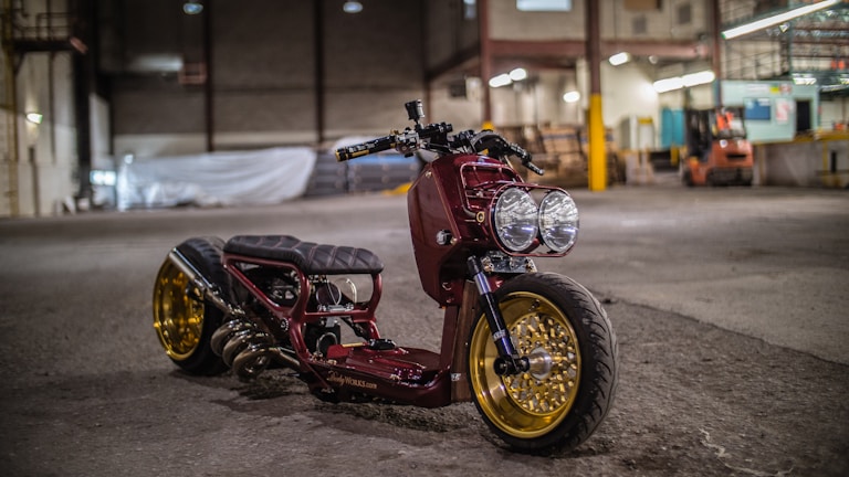 A custom motorcycle with a sleek design is parked inside an industrial warehouse. It features a maroon body, large dual headlights, and gold rims. The background includes high ceilings, concrete floors, and various equipment scattered around, giving a workshop-like atmosphere.