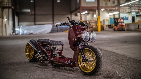 A custom motorcycle with a sleek design is parked inside an industrial warehouse. It features a maroon body, large dual headlights, and gold rims. The background includes high ceilings, concrete floors, and various equipment scattered around, giving a workshop-like atmosphere.
