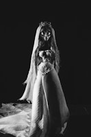 An animated character resembling a bride stands prominently in a dark setting. The character is wearing a long, flowing gown with intricate patterns and a veil. Its eyes are large and exaggerated, contributing to a dramatic, slightly haunting appearance.