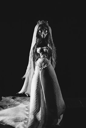 An animated character resembling a bride stands prominently in a dark setting. The character is wearing a long, flowing gown with intricate patterns and a veil. Its eyes are large and exaggerated, contributing to a dramatic, slightly haunting appearance.