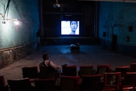 An individual is sitting alone in a dimly lit, old-style cinema theater with rows of empty red seats. A black-and-white film is being projected on a small screen at the front, displaying a close-up of a person's face. The walls are painted in a faded green color, and there are visible signs of wear and age.