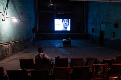 An individual is sitting alone in a dimly lit, old-style cinema theater with rows of empty red seats. A black-and-white film is being projected on a small screen at the front, displaying a close-up of a person's face. The walls are painted in a faded green color, and there are visible signs of wear and age.