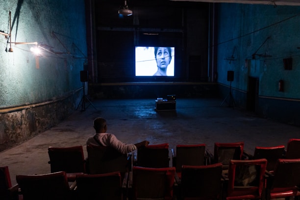 An individual is sitting alone in a dimly lit, old-style cinema theater with rows of empty red seats. A black-and-white film is being projected on a small screen at the front, displaying a close-up of a person's face. The walls are painted in a faded green color, and there are visible signs of wear and age.