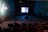 An individual is sitting alone in a dimly lit, old-style cinema theater with rows of empty red seats. A black-and-white film is being projected on a small screen at the front, displaying a close-up of a person's face. The walls are painted in a faded green color, and there are visible signs of wear and age.