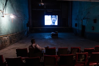 An individual is sitting alone in a dimly lit, old-style cinema theater with rows of empty red seats. A black-and-white film is being projected on a small screen at the front, displaying a close-up of a person's face. The walls are painted in a faded green color, and there are visible signs of wear and age.