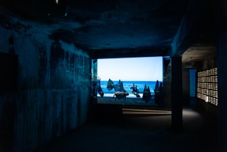 A dimly lit, narrow room with textured walls leads to a large window displaying a bright scene of a beach with scattered figures silhouetted against the sea. The contrast between the dark interior and the vibrant, sunlit outdoors creates a mysterious and immersive atmosphere.