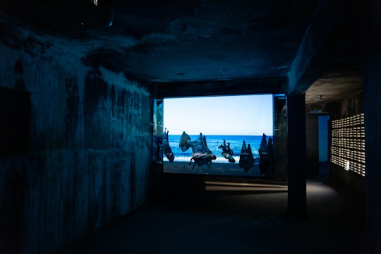 A dimly lit, narrow room with textured walls leads to a large window displaying a bright scene of a beach with scattered figures silhouetted against the sea. The contrast between the dark interior and the vibrant, sunlit outdoors creates a mysterious and immersive atmosphere.