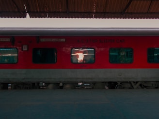 A red train carriage is stationary at a platform, under a canopy. The windows reveal the interior of the A.C. 3 tier sleeper car, and some text is visible on the exterior. The platform is empty and the train appears ready for boarding.