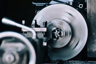 A close-up view of a spinning machine part, likely part of an industrial tool or lathe. The metal surface appears blurred due to motion, with surrounding mechanical components slightly out of focus.