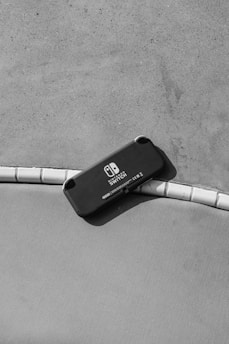 A black handheld gaming console lies on a curved surface, partially on tiles and partially on a fabric-like material. The branding on the console indicates it is a Nintendo Switch. Shadows are cast, suggesting directional lighting.