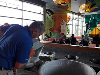 A man wearing a blue shirt and orange gloves is disposing of trash into a waste bin. The scene takes place in a cafeteria or dining area with modern decor and large windows. In the background, people are seated at tables, and there are colorful, artistic panels on the wall.