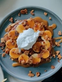 A round plate holds a dessert topped with banana slices, caramel sauce, crumbled cookies, and a dollop of whipped cream. The dish is presented on a simple blue ceramic plate, with the rich brown caramel and the yellow banana slices providing a contrast in colors.