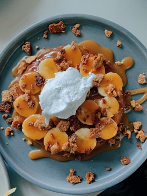 A round plate holds a dessert topped with banana slices, caramel sauce, crumbled cookies, and a dollop of whipped cream. The dish is presented on a simple blue ceramic plate, with the rich brown caramel and the yellow banana slices providing a contrast in colors.