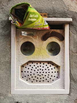 A mailbox embedded in a textured wall with a rolled-up newspaper sticking out from the slot. The mailbox has a rectangular shape with two circular button-like features and a perforated surface resembling a speaker grill. There is also a nameplate with text.