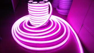 A coil of brightly glowing, flexible LED strip lights illuminated in bright pink, set against a darker background. The strips are neatly wound around a spool and extend outward in a spiral pattern.