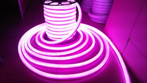 A coil of brightly glowing, flexible LED strip lights illuminated in bright pink, set against a darker background. The strips are neatly wound around a spool and extend outward in a spiral pattern.