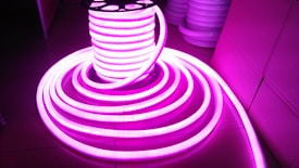 A coil of brightly glowing, flexible LED strip lights illuminated in bright pink, set against a darker background. The strips are neatly wound around a spool and extend outward in a spiral pattern.