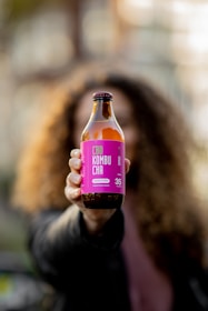 A person with long curly hair is holding up a bottle of CBD Kombucha with a vibrant pink label towards the camera. The background is blurred, focusing attention on the beverage.