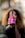 A person with long curly hair is holding up a bottle of CBD Kombucha with a vibrant pink label towards the camera. The background is blurred, focusing attention on the beverage.