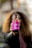 A person with long curly hair is holding up a bottle of CBD Kombucha with a vibrant pink label towards the camera. The background is blurred, focusing attention on the beverage.
